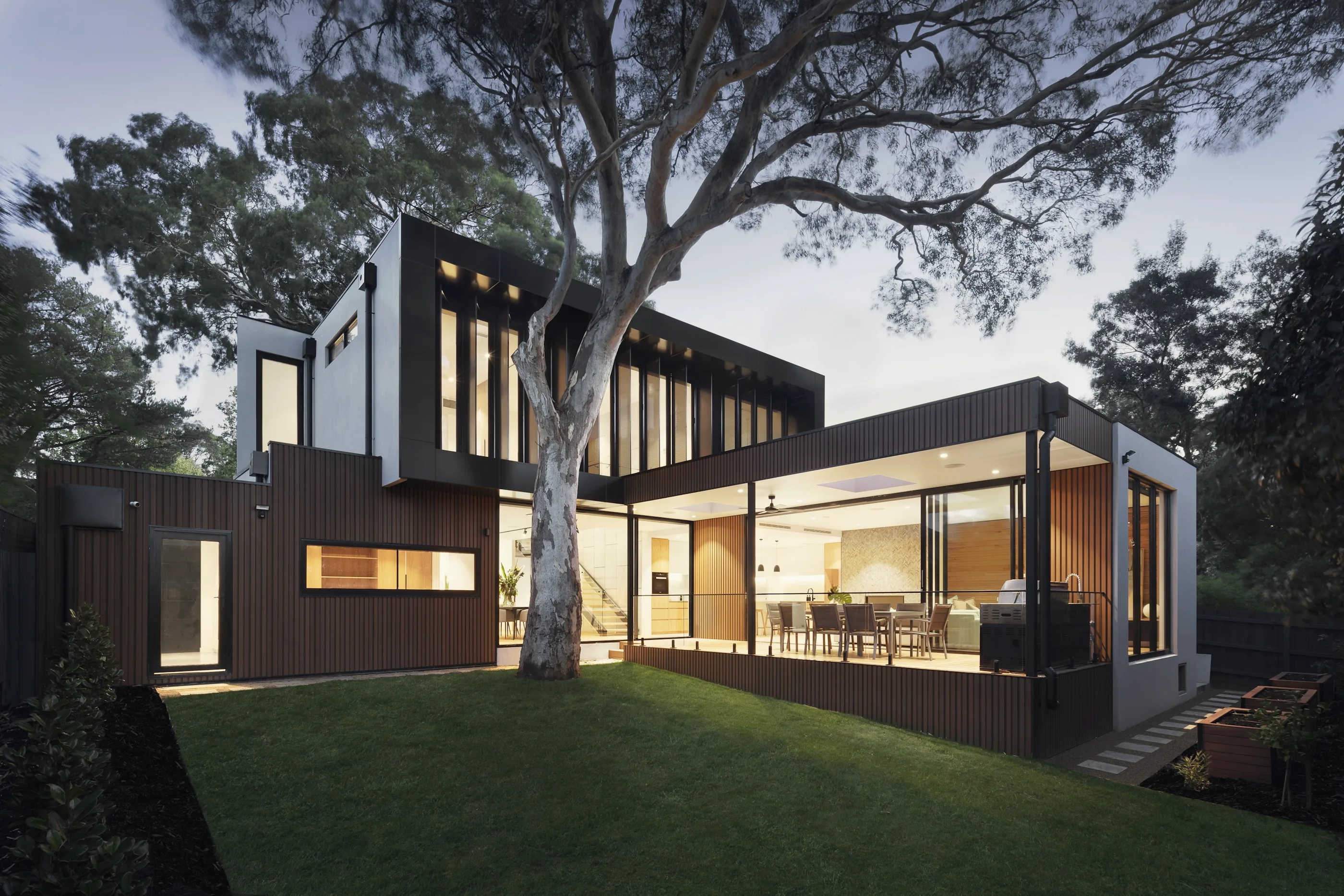 A low timber and glass residence glowing at dusk beneath a broad tree.
